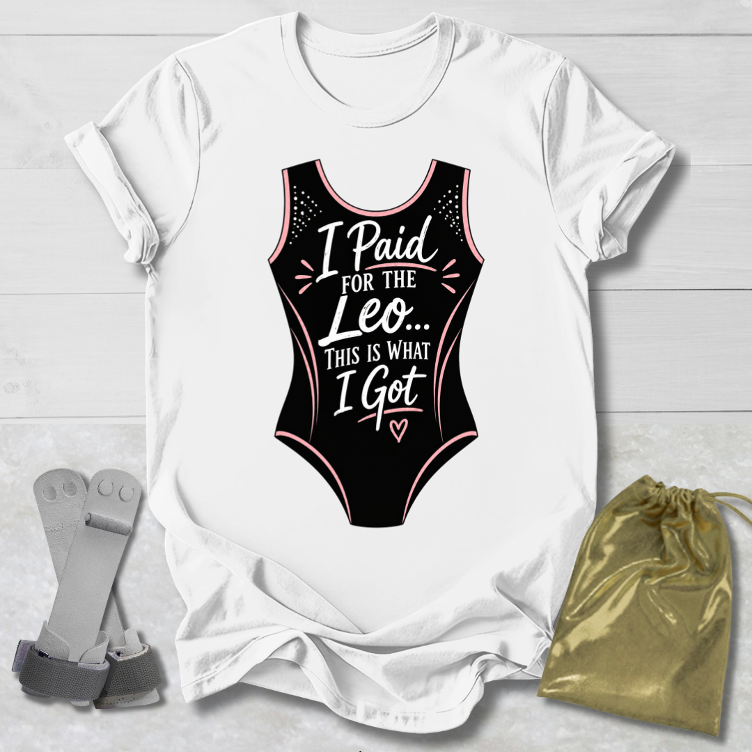 White t-shirt with black funny gymnastics leotard graphic and text, placed on a light wooden surface with socks and a drawstring bag.