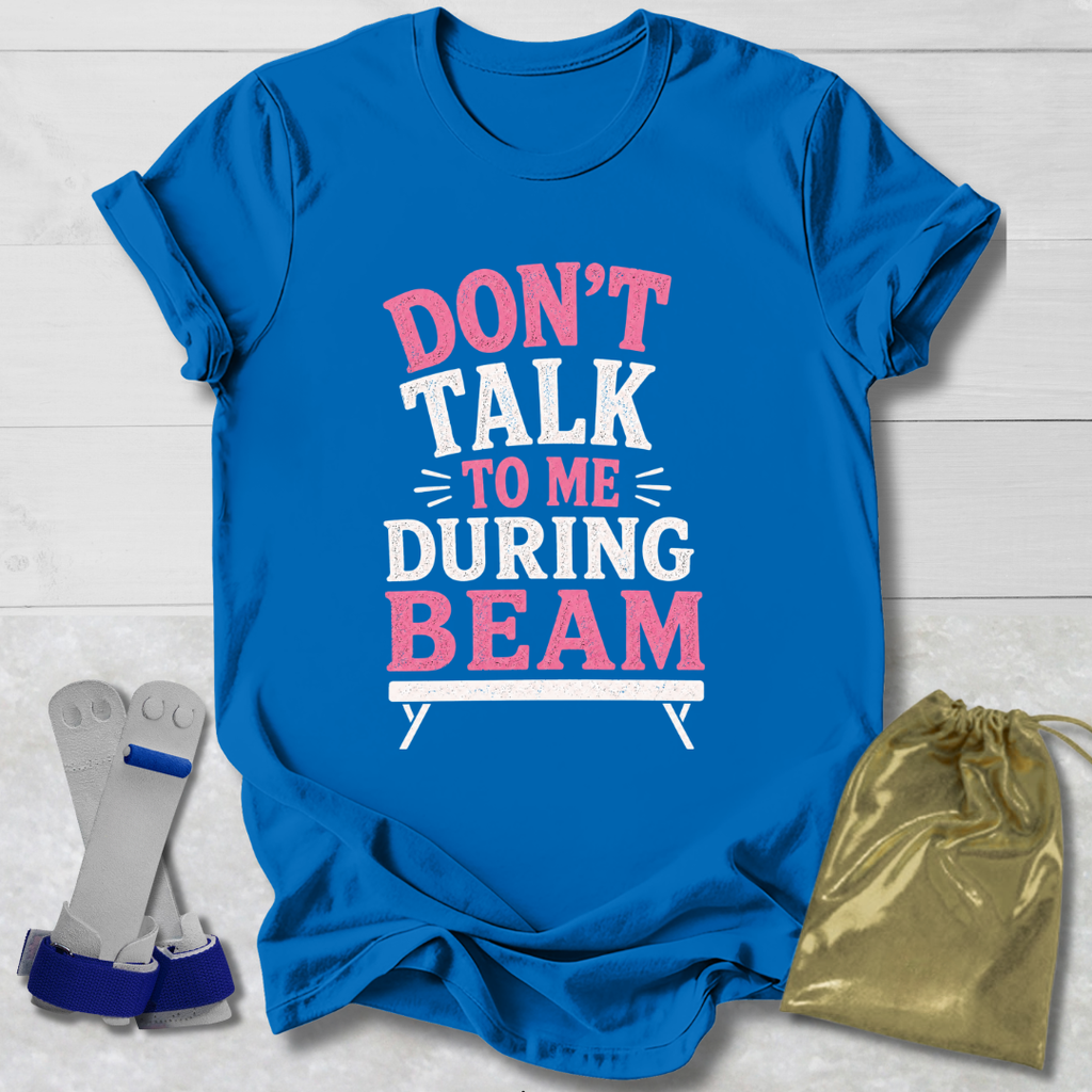 Blue t-shirt with 'Don't talk to me during beam' text on a light wooden surface, with socks and a drawstring bag in the foreground.