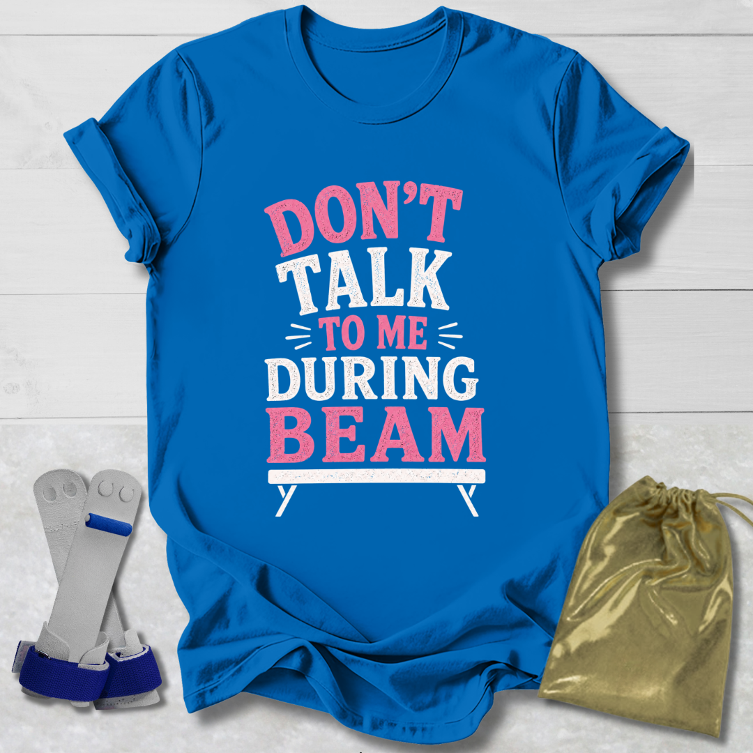 Blue t-shirt with 'Don't talk to me during beam' text on a light wooden surface, with socks and a drawstring bag in the foreground.