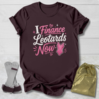 Maroon t-shirt with pink I finance leotards now gymnastics text and graphics on a light wooden surface, accompanied by socks and a drawstring bag.