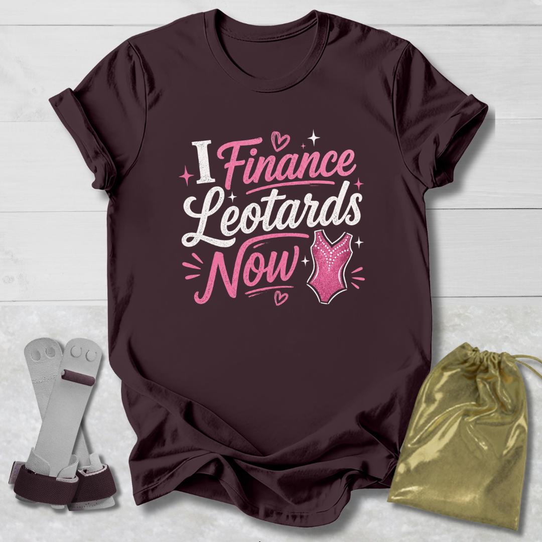 Maroon t-shirt with pink I finance leotards now gymnastics text and graphics on a light wooden surface, accompanied by socks and a drawstring bag.