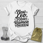 White t-shirt with 'New Leo Today, Ramen Tomorrow' text on a light wooden surface with socks and a drawstring bag.