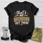 Black t-shirt with 'That's my grandson out there' text on a light wooden surface with socks and a drawstring bag.