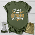 Green t-shirt with 'That's my grandson out there' text, socks, and drawstring bag on a light wooden surface.