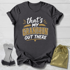 Black t-shirt with 'That's my grandson out there' text on a light wooden surface with socks and a drawstring bag.