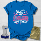 Blue t-shirt with text 'that's my granddaughter out there' on a light wooden surface, with socks and a drawstring bag in the foreground.