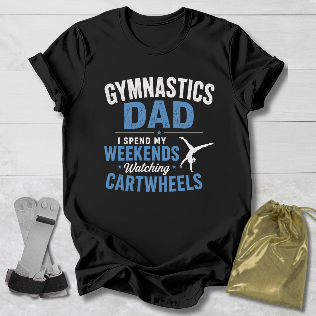 Black t-shirt with 'Gymnastics Dad' text on a wooden surface with socks and a drawstring bag.