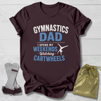 Maroon t-shirt with 'Gymnastics Dad' text on a light wooden surface with socks and a drawstring bag.