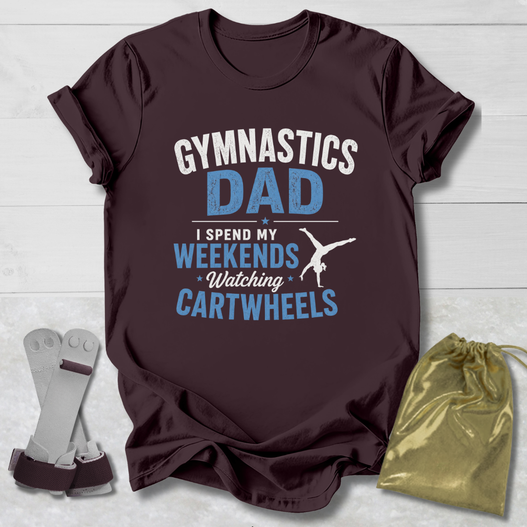 Maroon t-shirt with 'Gymnastics Dad' text on a light wooden surface with socks and a drawstring bag.