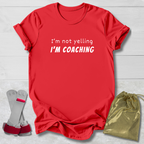 Red t-shirt with 'I'm not yelling I'm coaching' text, socks, and drawstring bag on a light wooden surface.