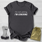 Dark gray t-shirt with 'I'm not yelling I'm coaching' text on a light wooden surface with socks and a drawstring bag.