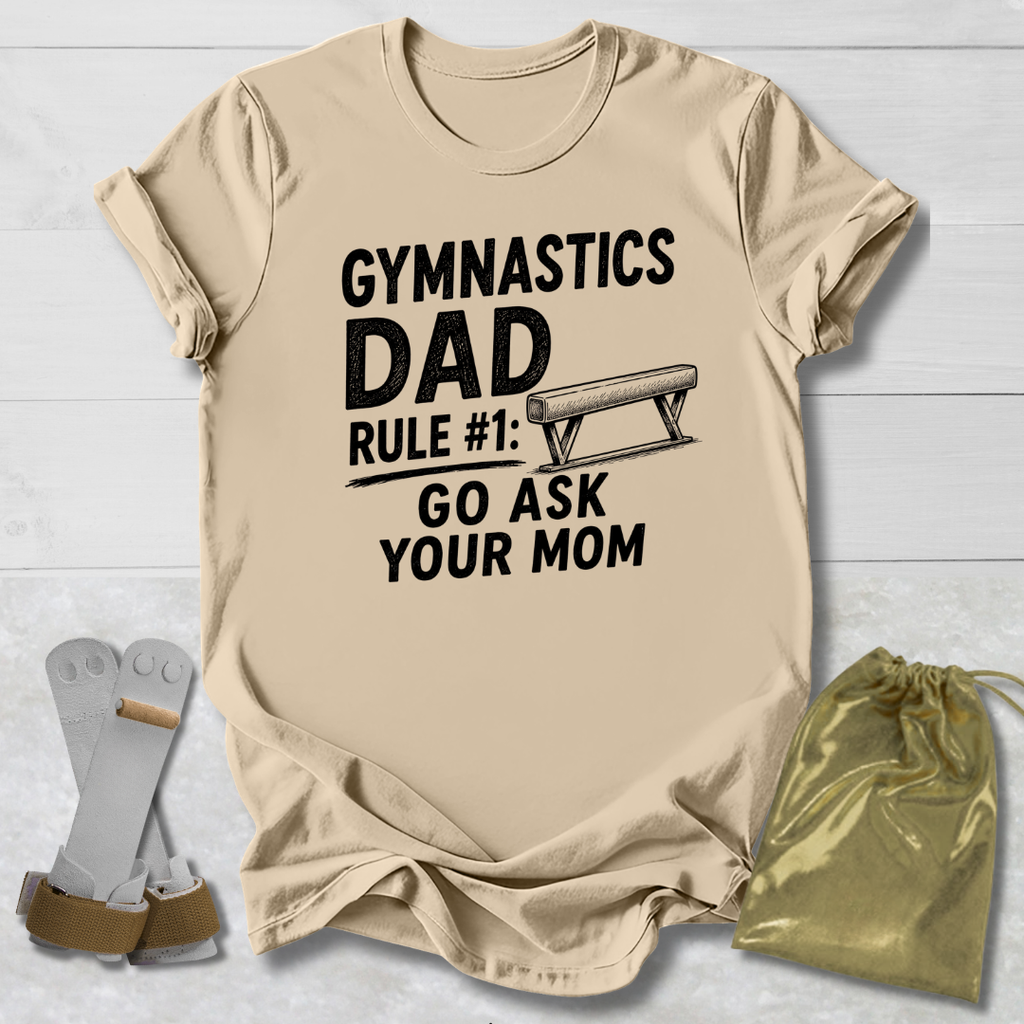 Beige t-shirt with 'Gymnastics Dad' text on a light wooden surface with socks and a drawstring bag.