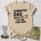 Beige t-shirt with 'Gymnastics Dad' text on a light wooden surface with socks and a drawstring bag.