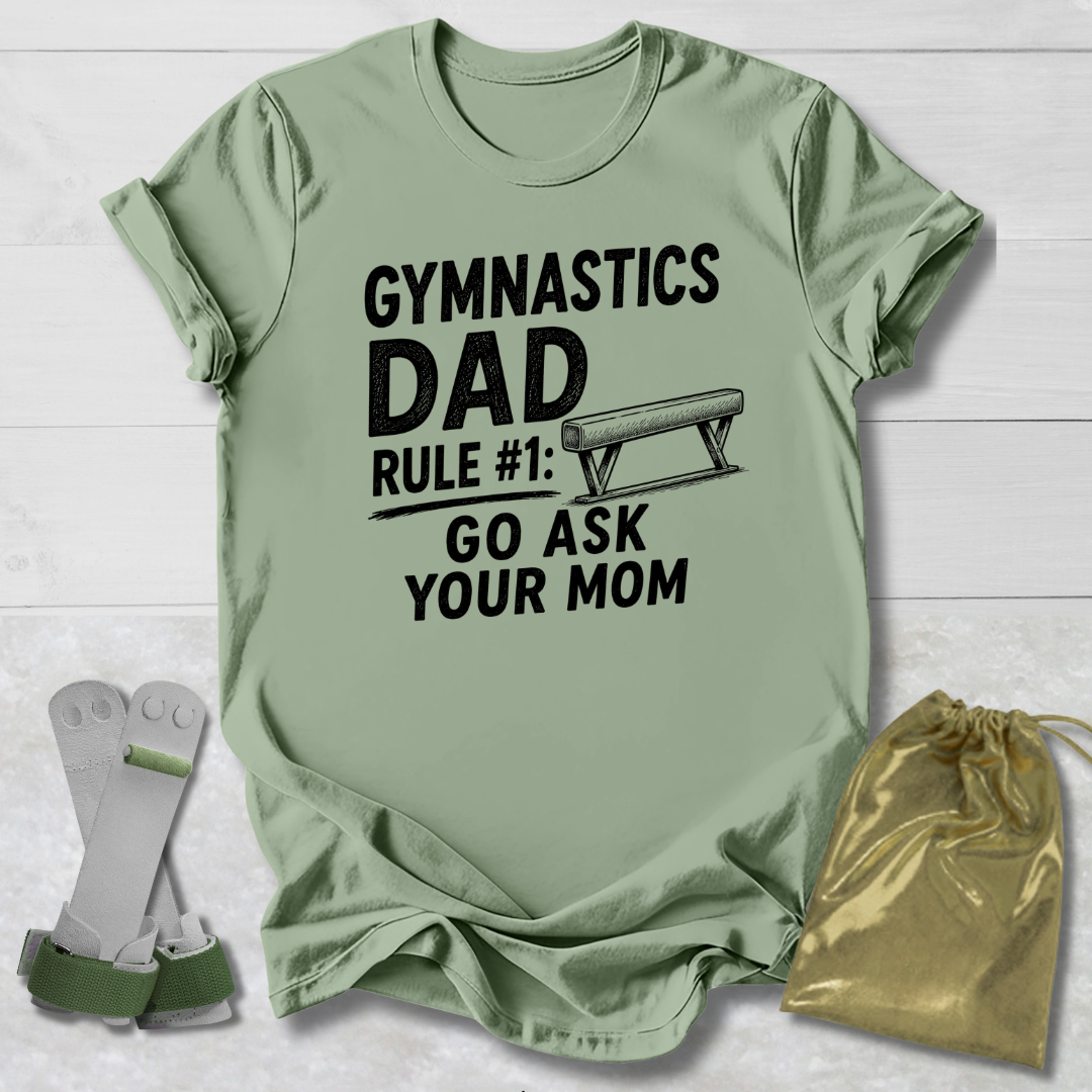Green t-shirt with 'Gymnastics Dad' text on a wooden surface with socks and a drawstring bag.