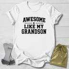 White t-shirt with 'Awesome Like My Grandson' text on a light wooden surface with socks and a drawstring bag.