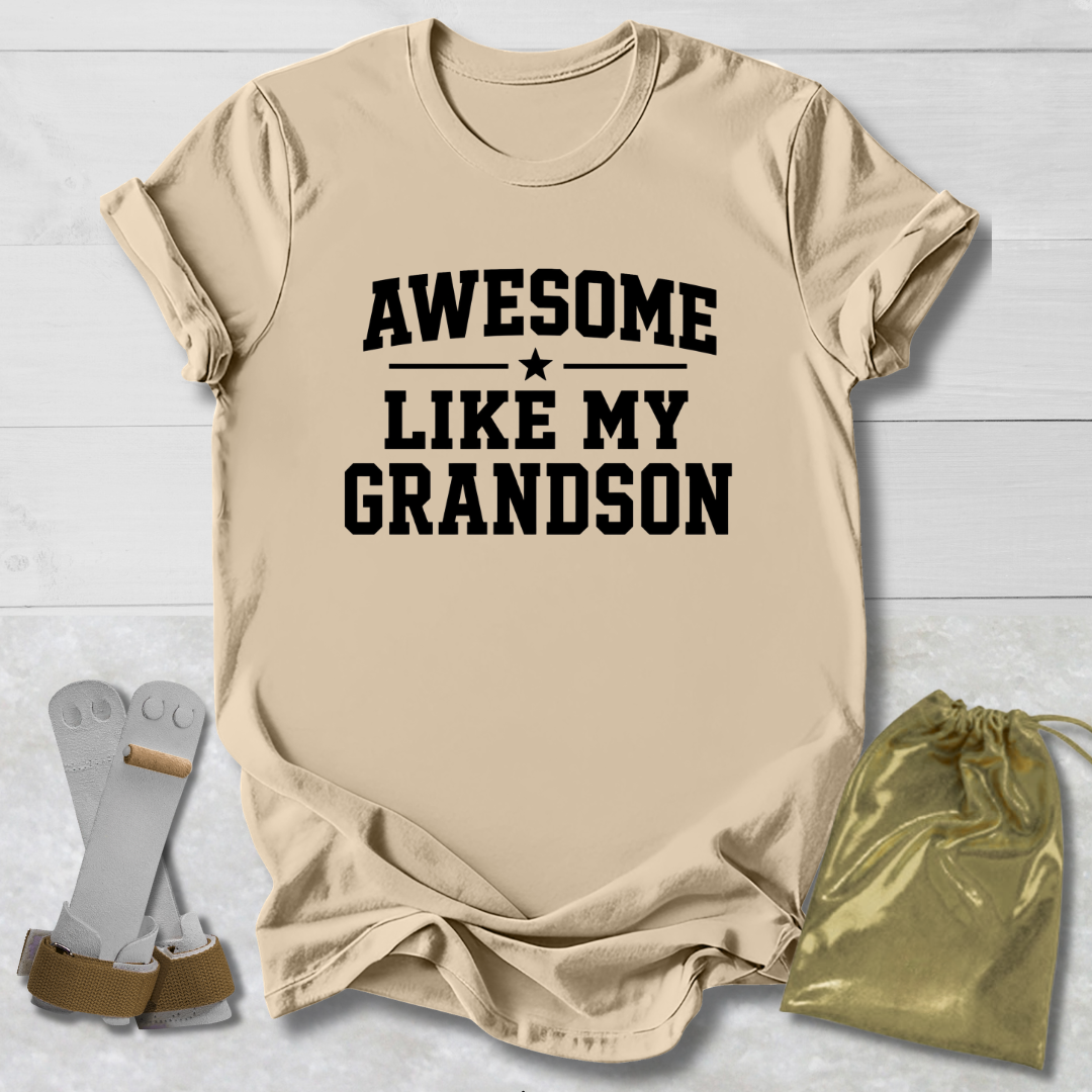 Beige t-shirt with 'Awesome Like My Grandson' text on a light wooden surface with socks and a drawstring bag.