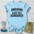 Light blue t-shirt with 'Awesome like my granddaughter' text, socks, and drawstring bag on a light wooden surface.