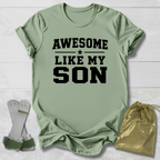 Green t-shirt with 'Awesome Like My Son' text, socks, and drawstring bag on a light wooden surface.
