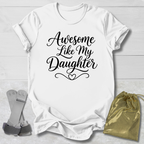 White t-shirt with 'Awesome Like My Daughter' text on a light wooden surface with socks and a drawstring bag.