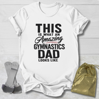 White t-shirt with text 'This is what an amazing gymnastics dad looks like' on a light wooden surface.