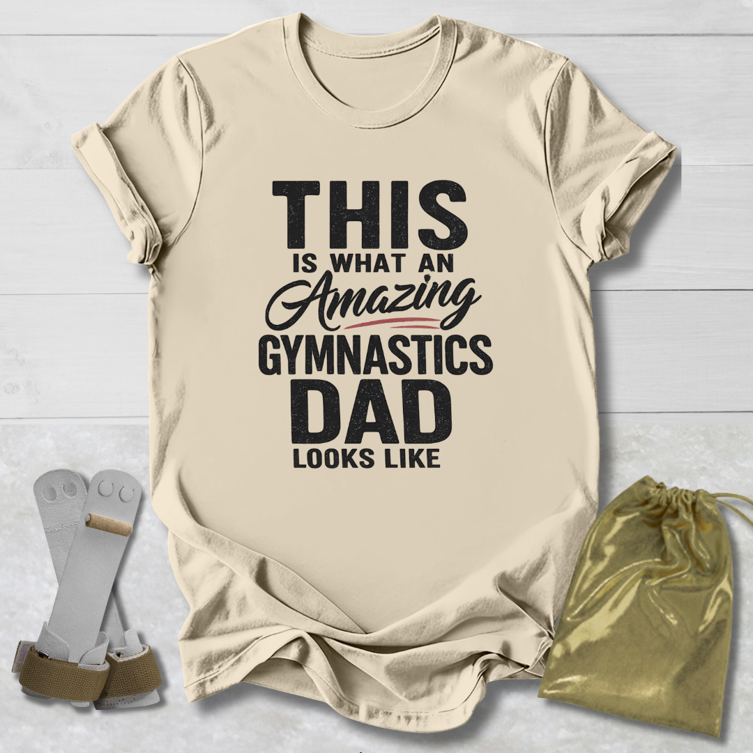 Beige t-shirt with text 'This is what an amazing gymnastics dad looks like' on a light wooden surface.
