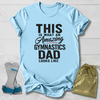 Light blue t-shirt with text 'This is what an amazing gymnastics dad looks like' on a wooden surface, with socks and a drawstring bag.