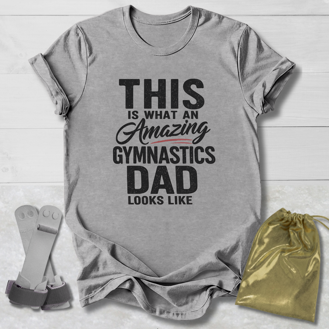 Gray t-shirt with text 'This is what an amazing gymnastics dad looks like' on a wooden surface with socks and a drawstring bag.