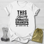 White t-shirt with black text 'This is what an amazing gymnastics grandpa looks like' on a light wooden surface.