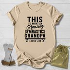 Beige t-shirt with black text 'This is what an amazing gymnastics grandpa looks like' on a light wooden surface.