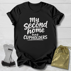 Black t-shirt with 'My second home has cupholders' text on a light wooden surface with socks and a drawstring bag.