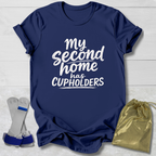 Navy blue t-shirt with white text 'My second home has cupholders' on a light wooden surface with socks and a drawstring bag.