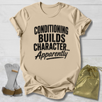 Beige t-shirt with text 'Conditioning Builds Character... Apparently' on a light wooden surface.