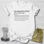 White t-shirt with 'Gymnastics Dad' definition text on a wooden surface with socks and a drawstring bag.