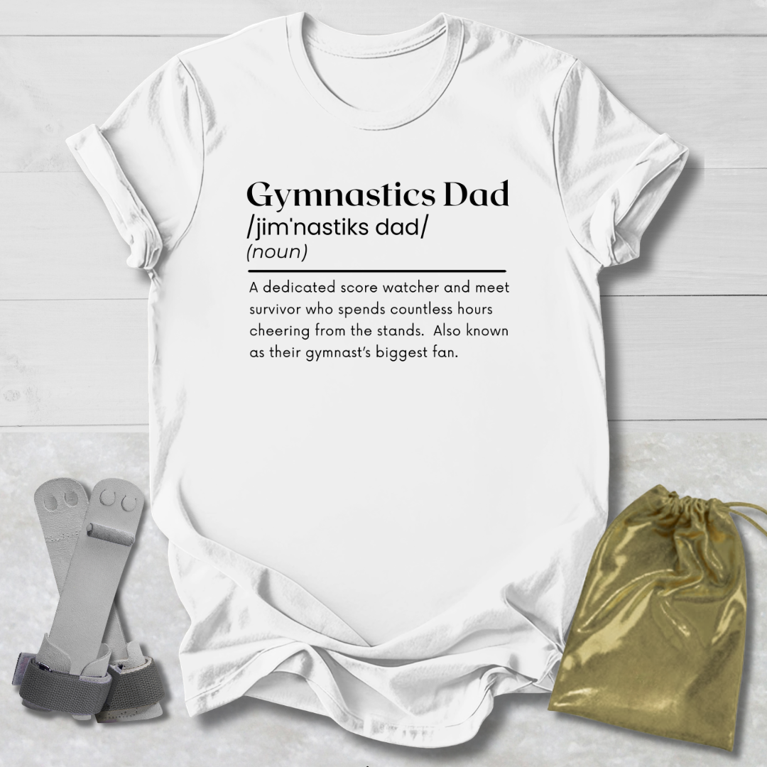 White t-shirt with 'Gymnastics Dad' definition text on a wooden surface with socks and a drawstring bag.
