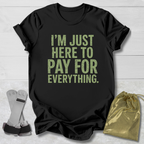 Black t-shirt with yellow text 'I'm just here to pay for everything' on a light wooden surface.