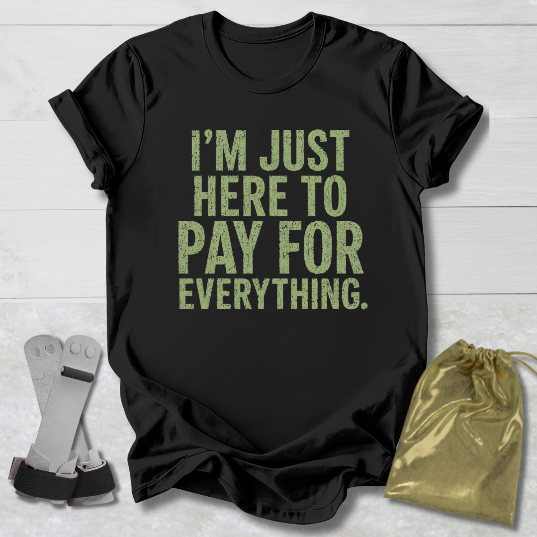 Black t-shirt with yellow text 'I'm just here to pay for everything' on a light wooden surface.
