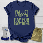 Navy blue t-shirt with yellow text 'I'm just here to pay for everything' on a light wooden surface.