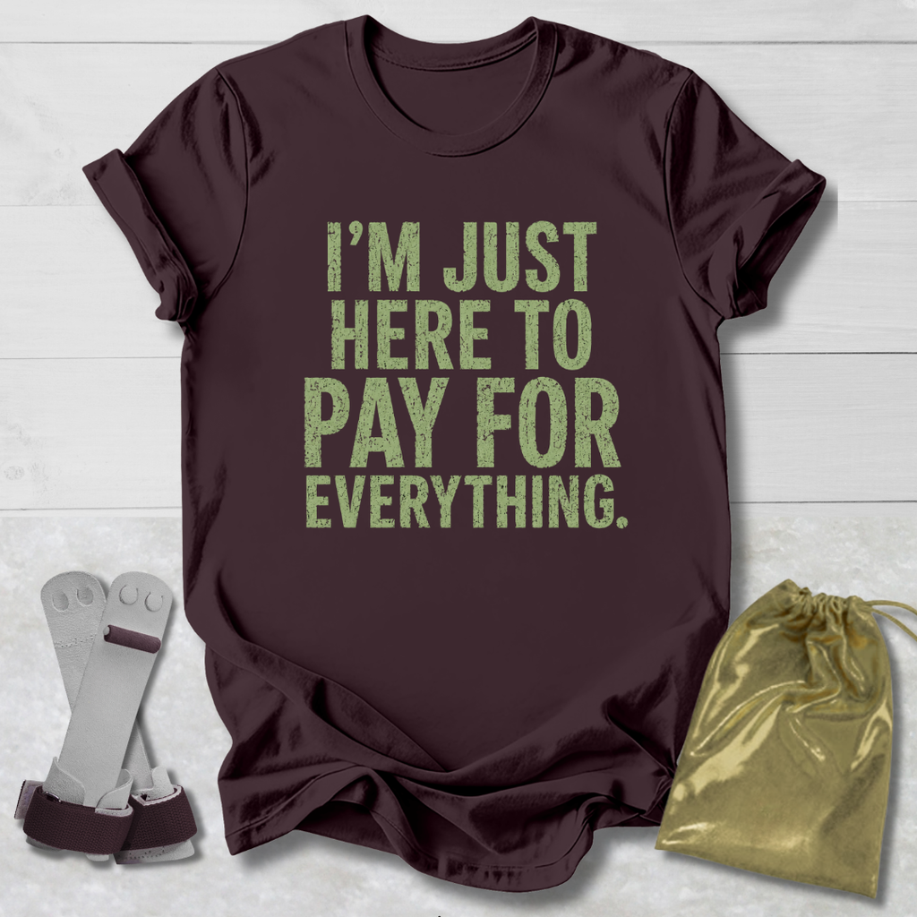 Maroon t-shirt with green text 'I'm just here to pay for everything' on a light wooden surface.