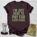 Maroon t-shirt with green text 'I'm just here to pay for everything' on a light wooden surface.
