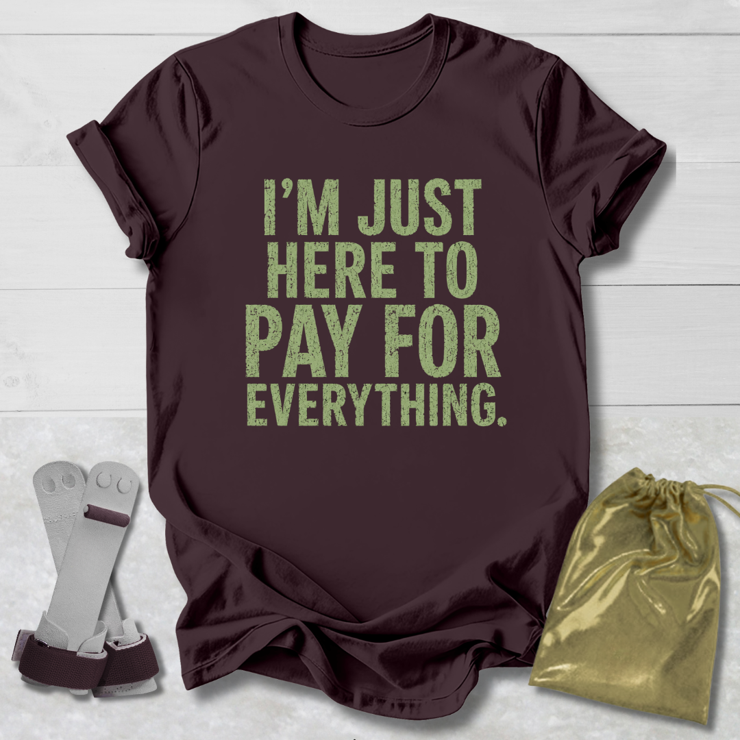 Maroon t-shirt with green text 'I'm just here to pay for everything' on a light wooden surface.