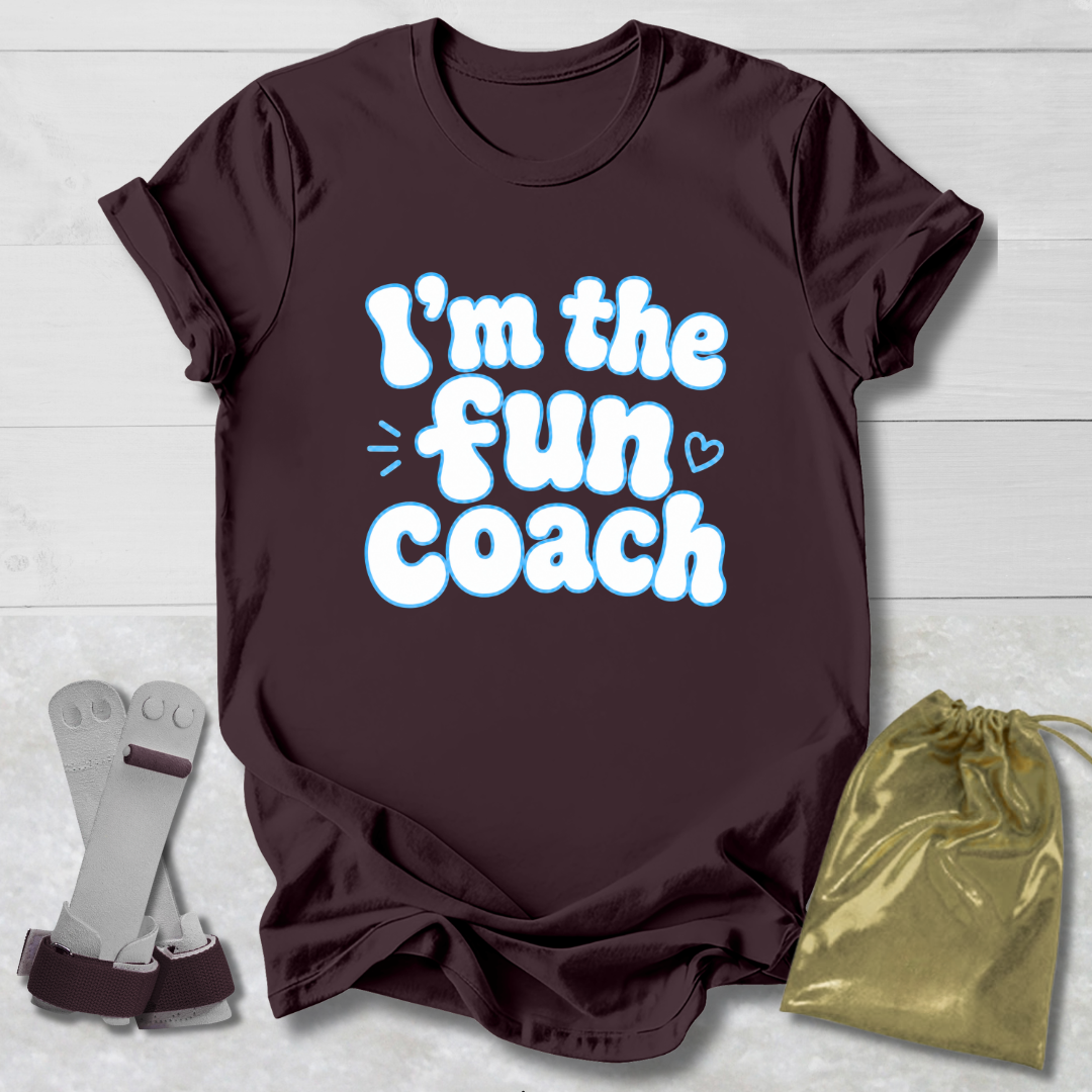 Maroon t-shirt with 'I'm the fun coach' text on a light wooden surface, with socks and a drawstring bag in the foreground.