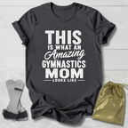 Black t-shirt with 'Amazing Gymnastics Mom' text on a light wooden surface with socks and a drawstring bag.