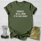 Green t-shirt with text 'Seriously... We all know I'm the coach's favorite' on a light wooden surface.