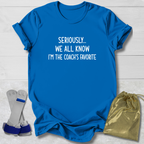 Blue t-shirt with text 'Seriously... We all know I'm the coach's favorite' on a light wooden surface.