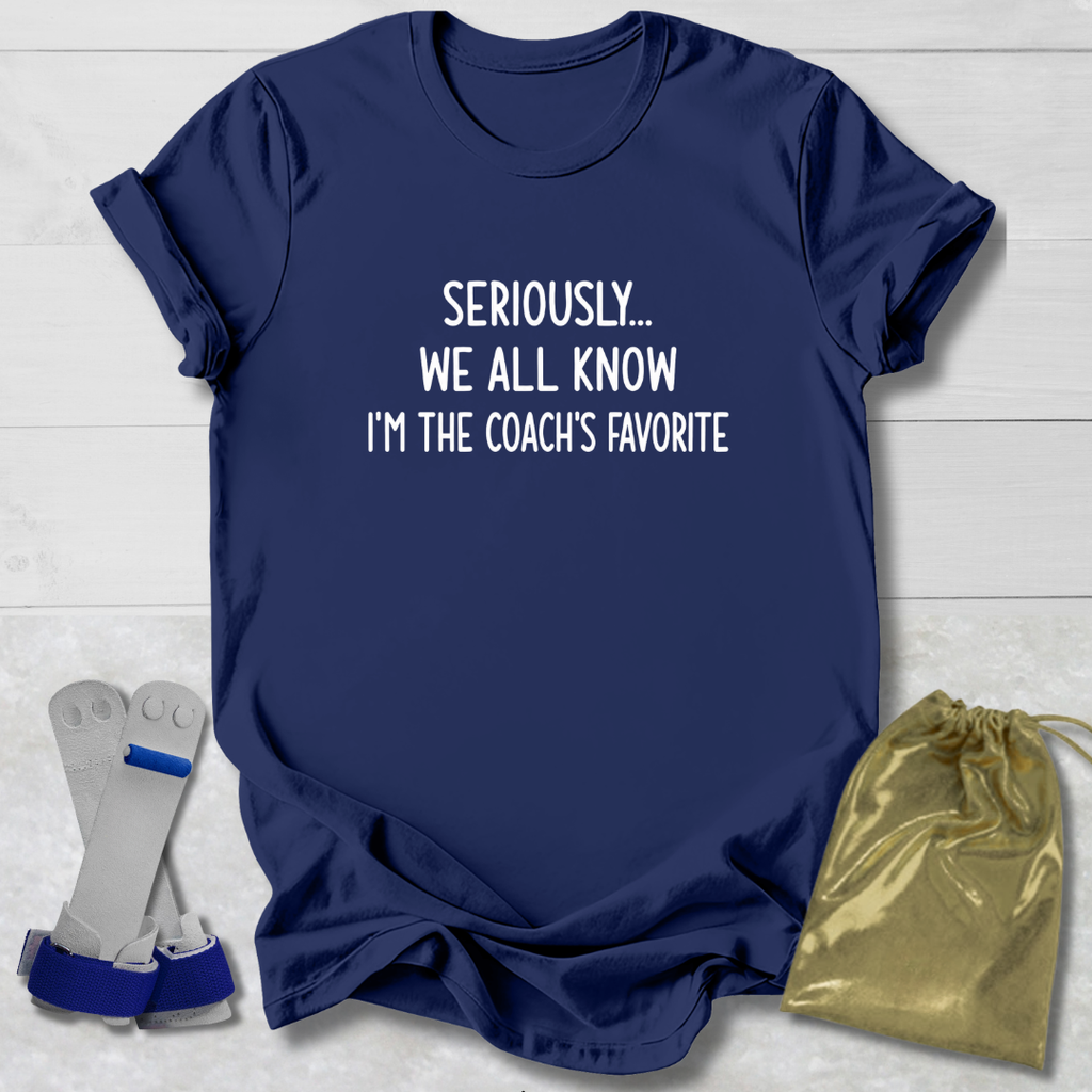Navy blue t-shirt with text 'Seriously... We all know I'm the coach's favorite' on a light wooden surface.