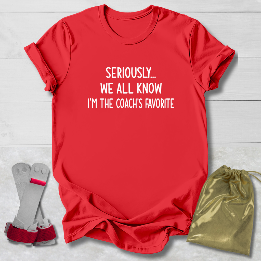 Red t-shirt with text 'Seriously... We all know I'm the coach's favorite' on a light wooden surface.