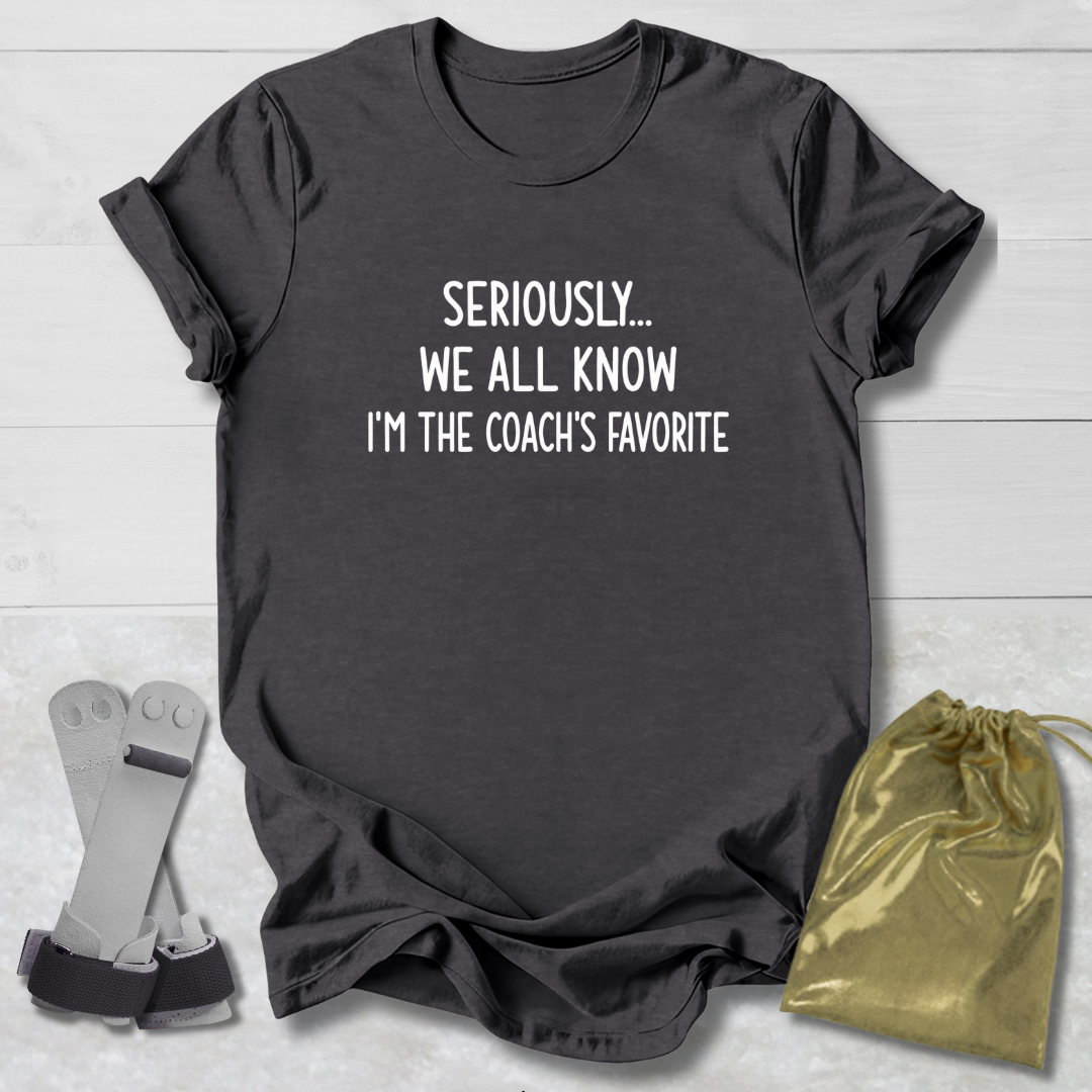 Black t-shirt with text 'Seriously... We all know I'm the coach's favorite' on a light wooden surface.