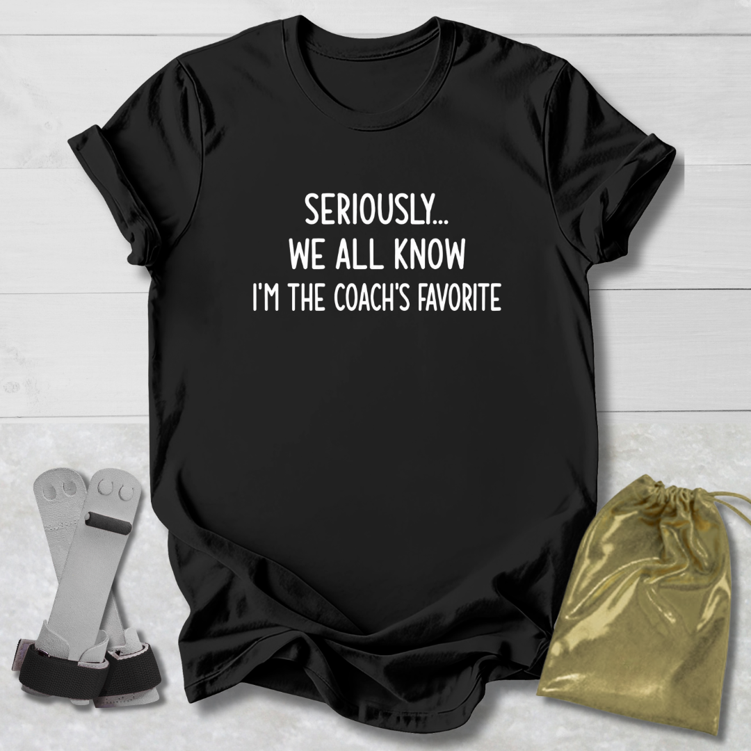 Black t-shirt with text 'Seriously... We all know I'm the coach's favorite' on a light wooden surface.