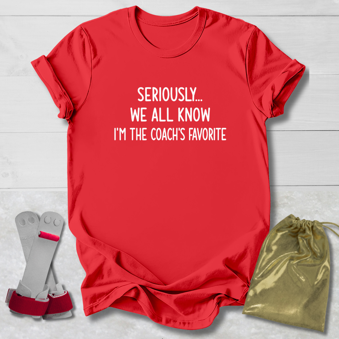 Red t-shirt with text 'Seriously... We all know I'm the coach's favorite' on a light wooden surface.