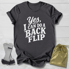 Black t-shirt with 'Yes, I can do a back flip' text on a light wooden surface with socks and a drawstring bag.